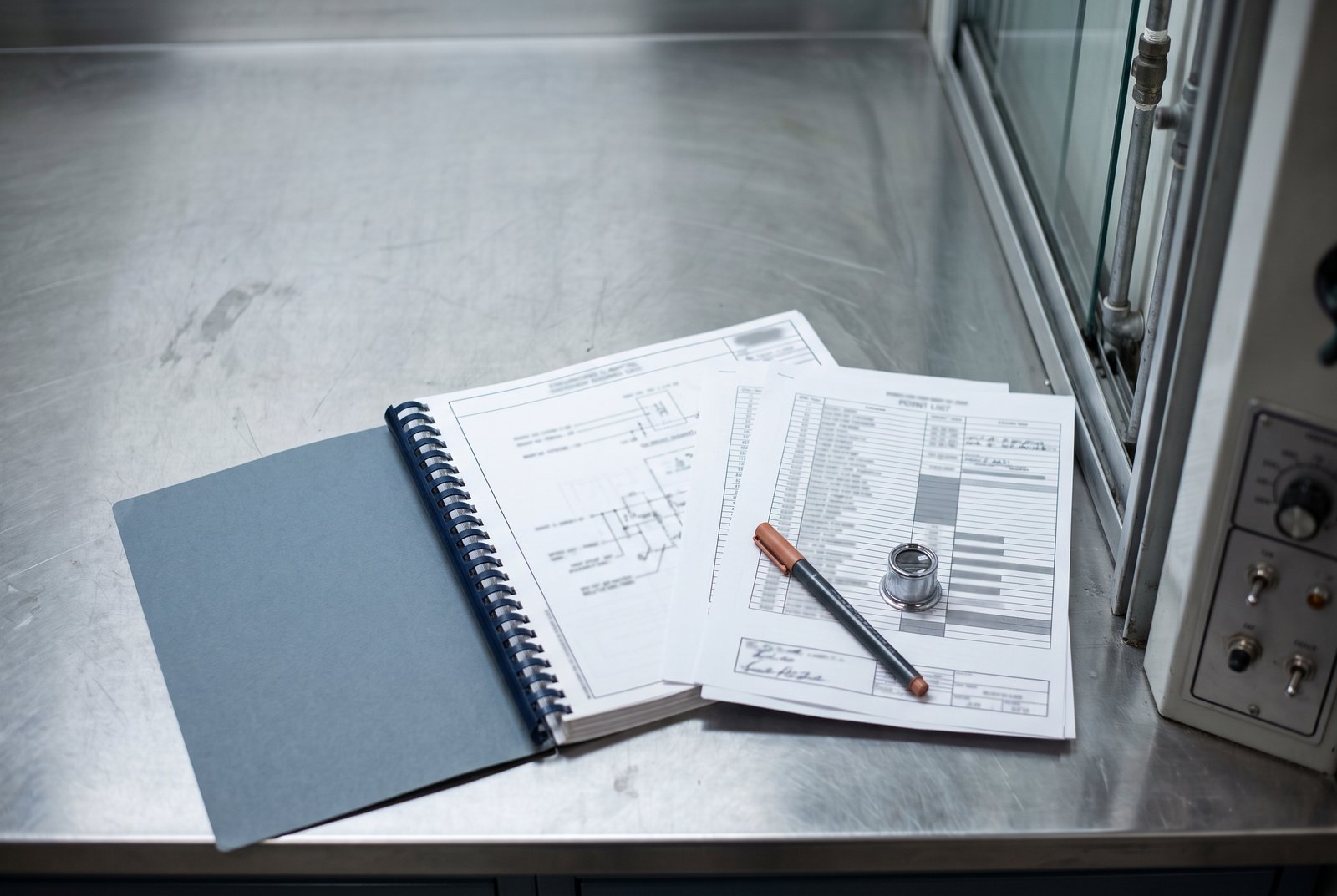 A bound laboratory submittal package and point list on a stainless bench beside a fume hood and a control panel.